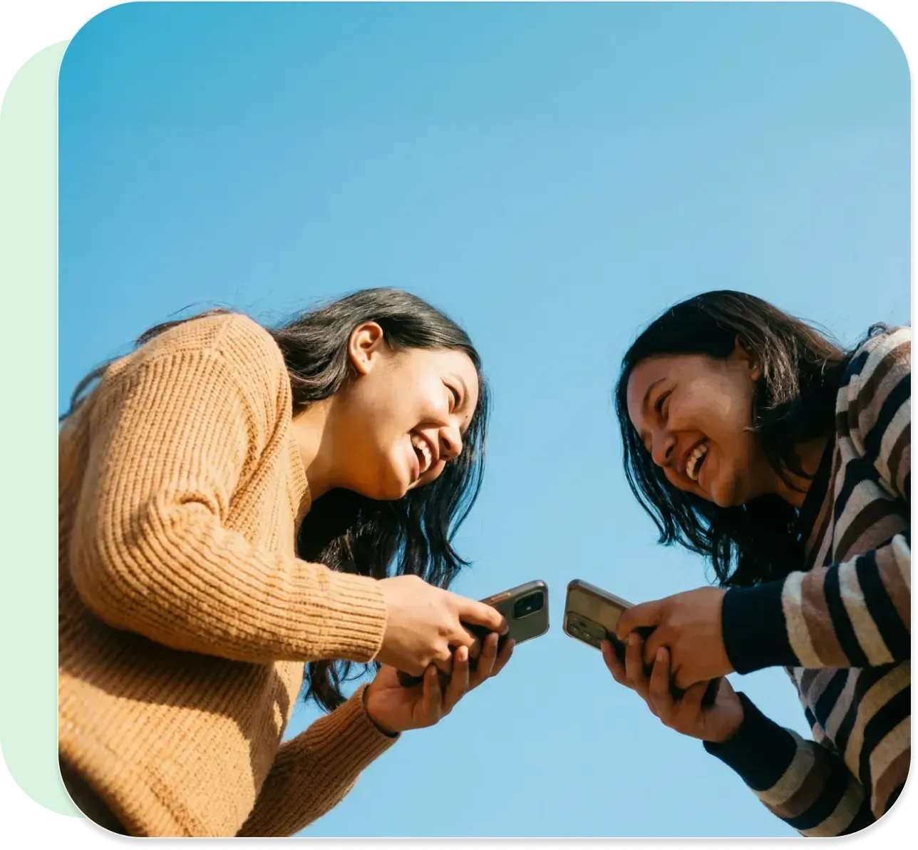 Two friends smiling while using phones outdoors